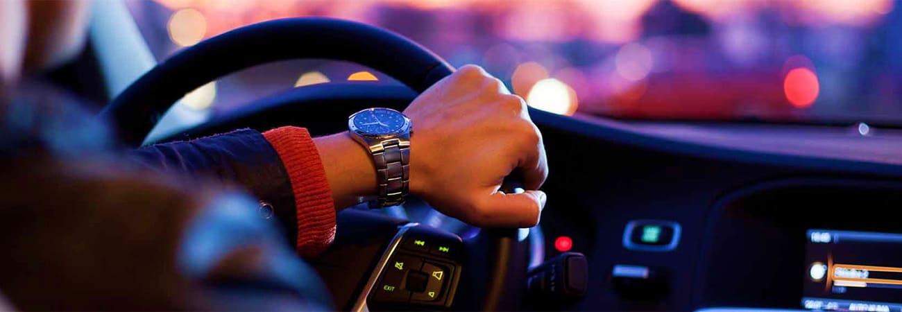 Jeddah Airport Pick Up Driver's hand on steering wheel wearing luxury watch, car interior at dusk with city lights in background